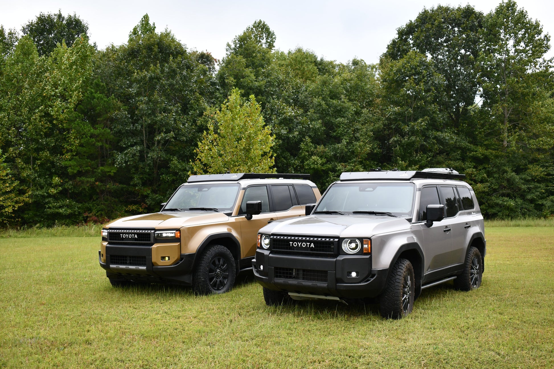 Two 2024+ Toyota Land Cruiser SUVs, one tan and one gray, are parked on grass with trees behind. Both feature rugged designs and are equipped with Ruggedized Aluminum Low Profile Racks for the 250 Series on their roof racks, facing slightly left.