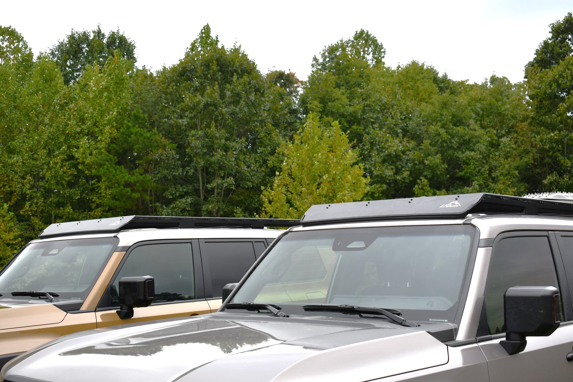 Two 2024+ 250 Series Land Cruisers equipped with Ruggedized Aluminum Low Profile Racks are parked side by side under an overcast sky, their tan and silver exteriors set against green trees and ready for adventure.