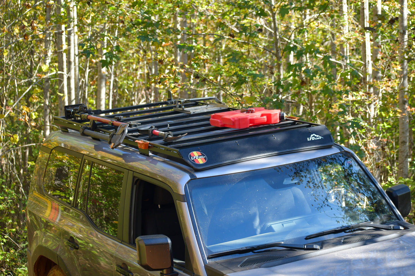 A gray SUV in a wooded area has a Ruggedized Aluminum Low Profile Rack for the 2024+ 250 Series Land Cruiser, with modular crossbars holding tools, a red fuel can, and camping gear. Sunlight streams through green trees in the background.