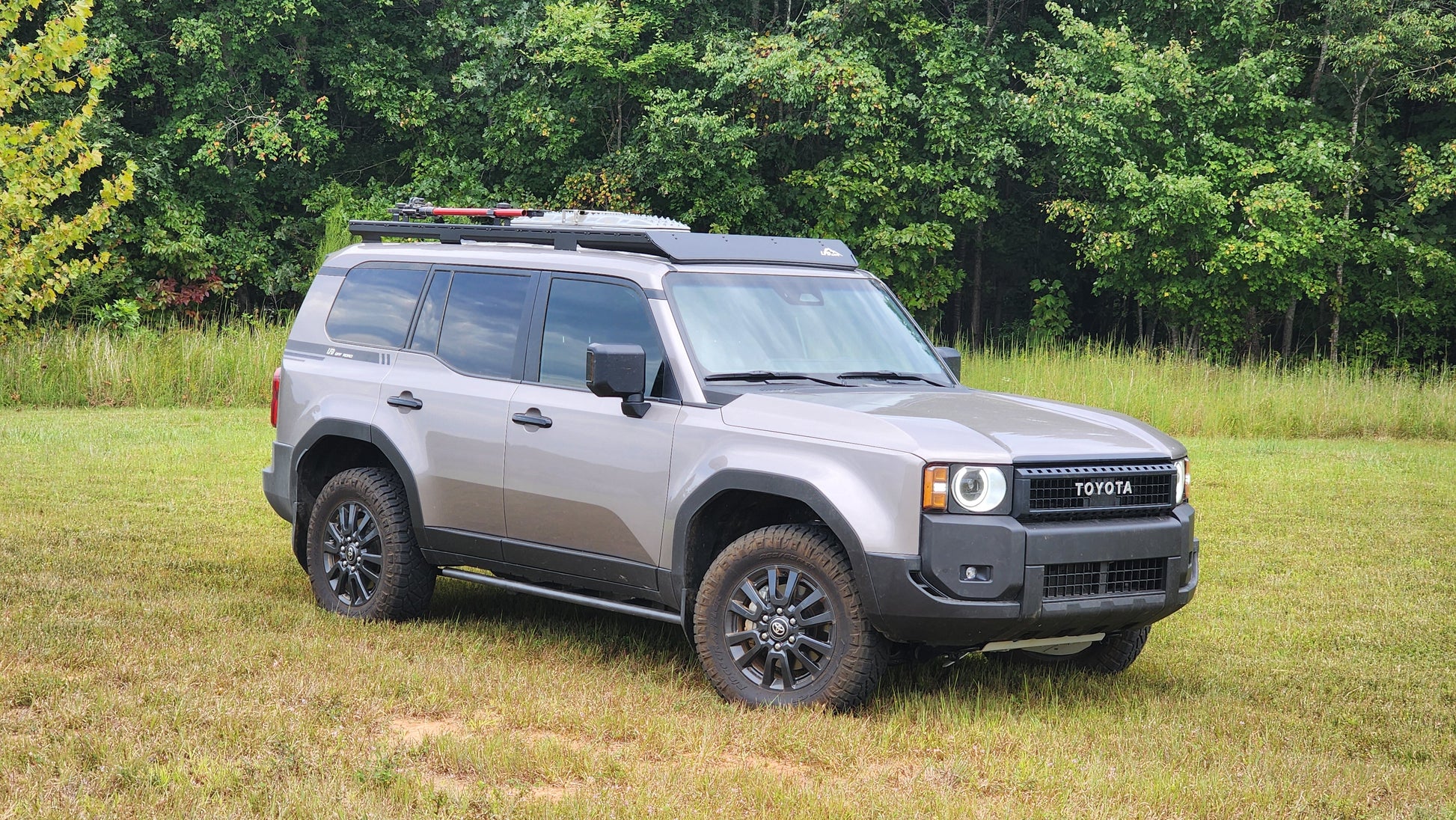 A silver 2024 Toyota Land Cruiser is parked on grass before dense green trees, fitted with a Ruggedized Aluminum Low Profile Rack - 250 Series Land Cruiser (2024+) and black wheels.