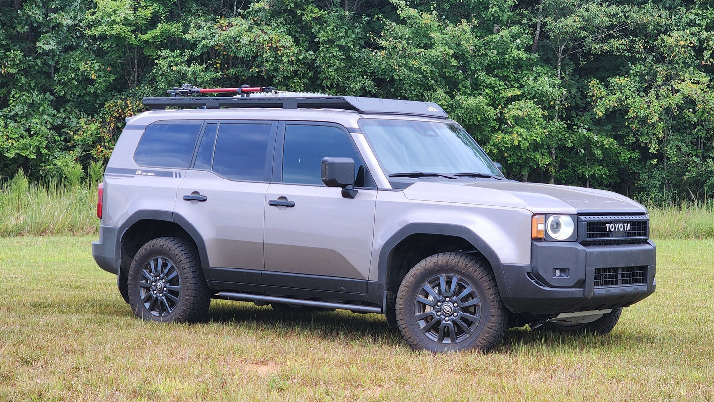 A gray 2024+ Toyota Land Cruiser 250 Series SUV with black wheels and a Ruggedized Aluminum Low Profile Rack is parked on grass in front of dense green trees.