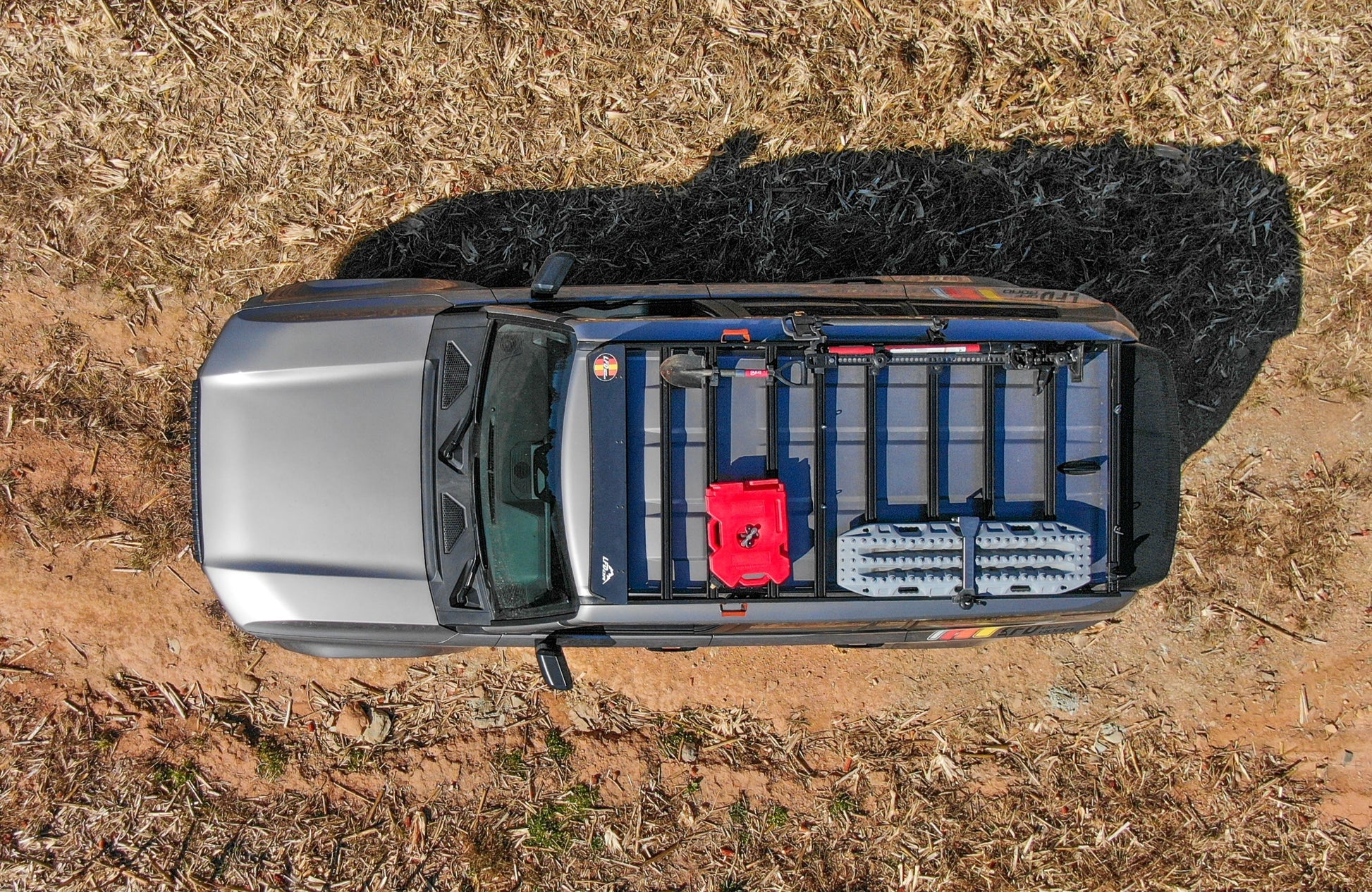 Top-down view of a gray pickup on dry grass, its bed organized with a red fuel can, traction boards, and tools using modular crossbars from the Ruggedized Aluminum Low Profile Rack - 250 Series Land Cruiser (2024+).