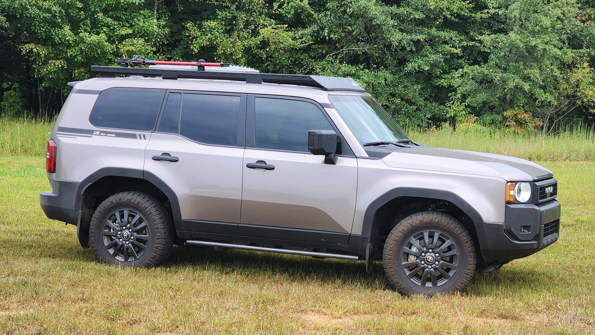 A gray SUV with black trim and off-road tires is parked on grass before dense green trees, featuring a Ruggedized Aluminum Low Profile Rack designed for the 2024+ 250 Series Land Cruiser, loaded with equipment on top.