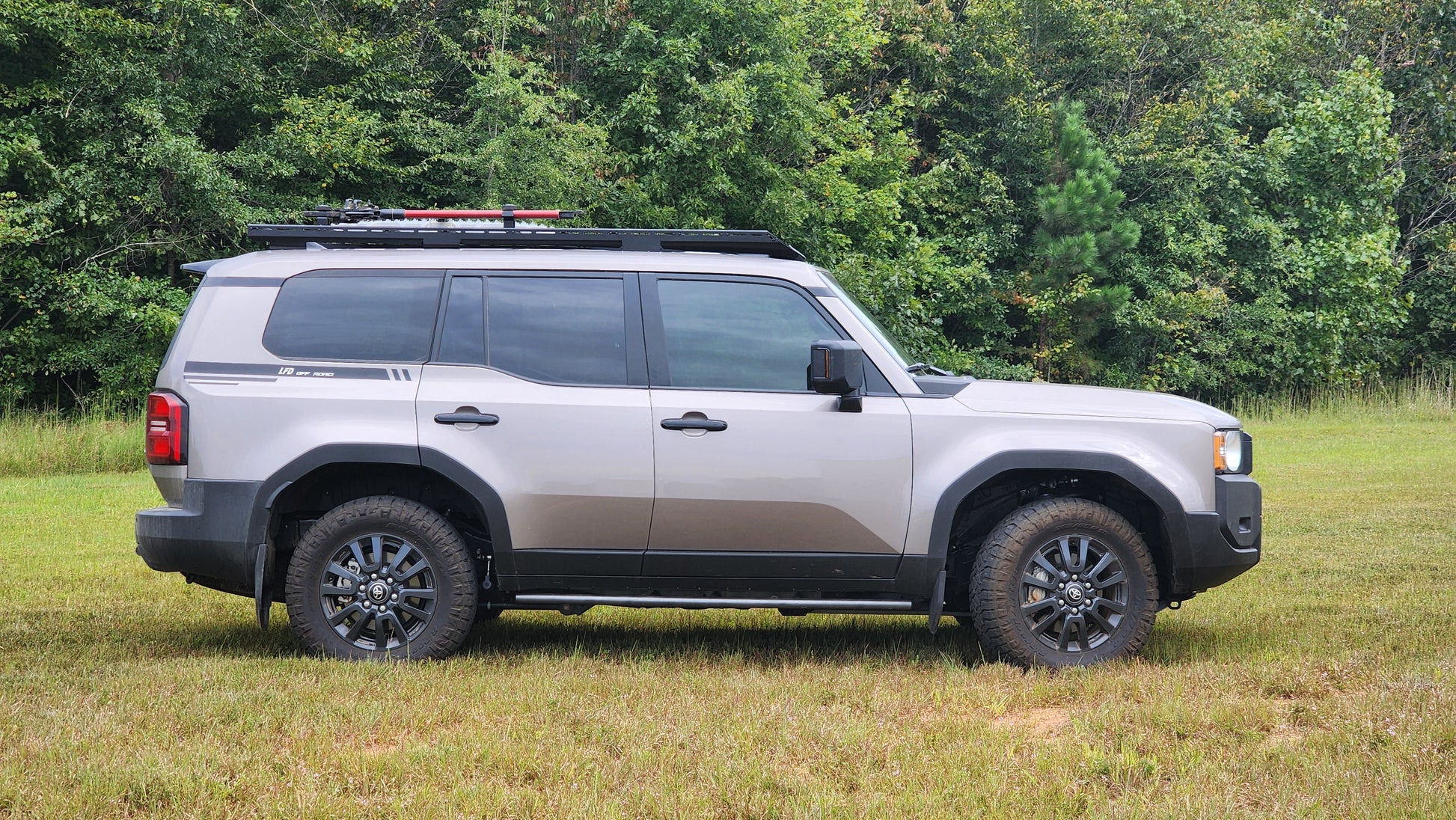 A gray SUV with black wheels and a Ruggedized Aluminum Low Profile Rack - 250 Series Land Cruiser (2024+) is parked on grass before dense green trees, seen from the side.
