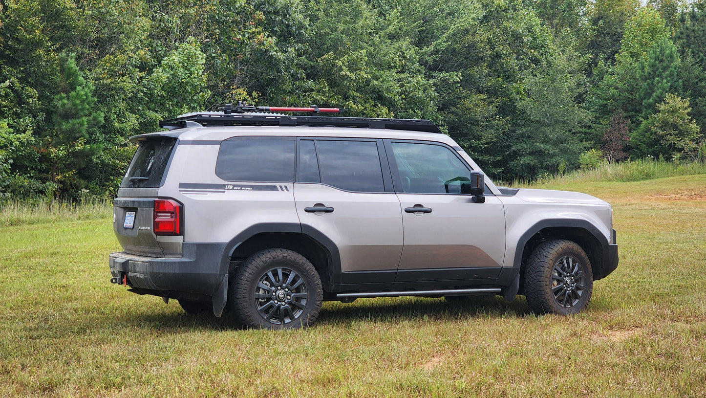 A silver 2024+ Land Cruiser 250 Series with black accents and off-road tires sits on grassy terrain amid dense foliage, equipped with a Ruggedized Aluminum Low Profile Rack for outdoor adventures.