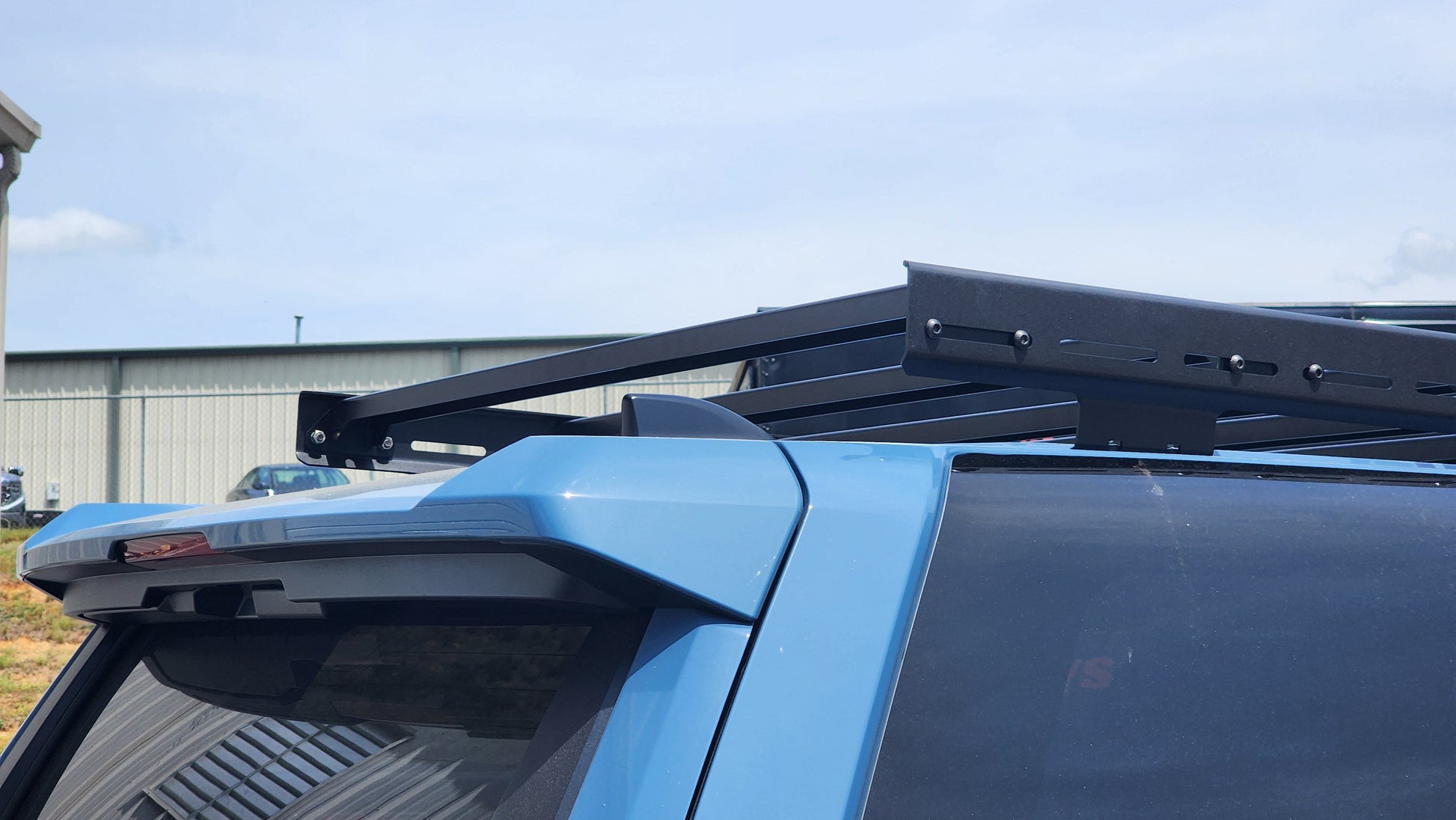 Close-up of a blue 6th Gen 4Runner featuring the Ruggedized Low Profile Roof Rack, parked outdoors near a light-colored building under a partly cloudy sky.