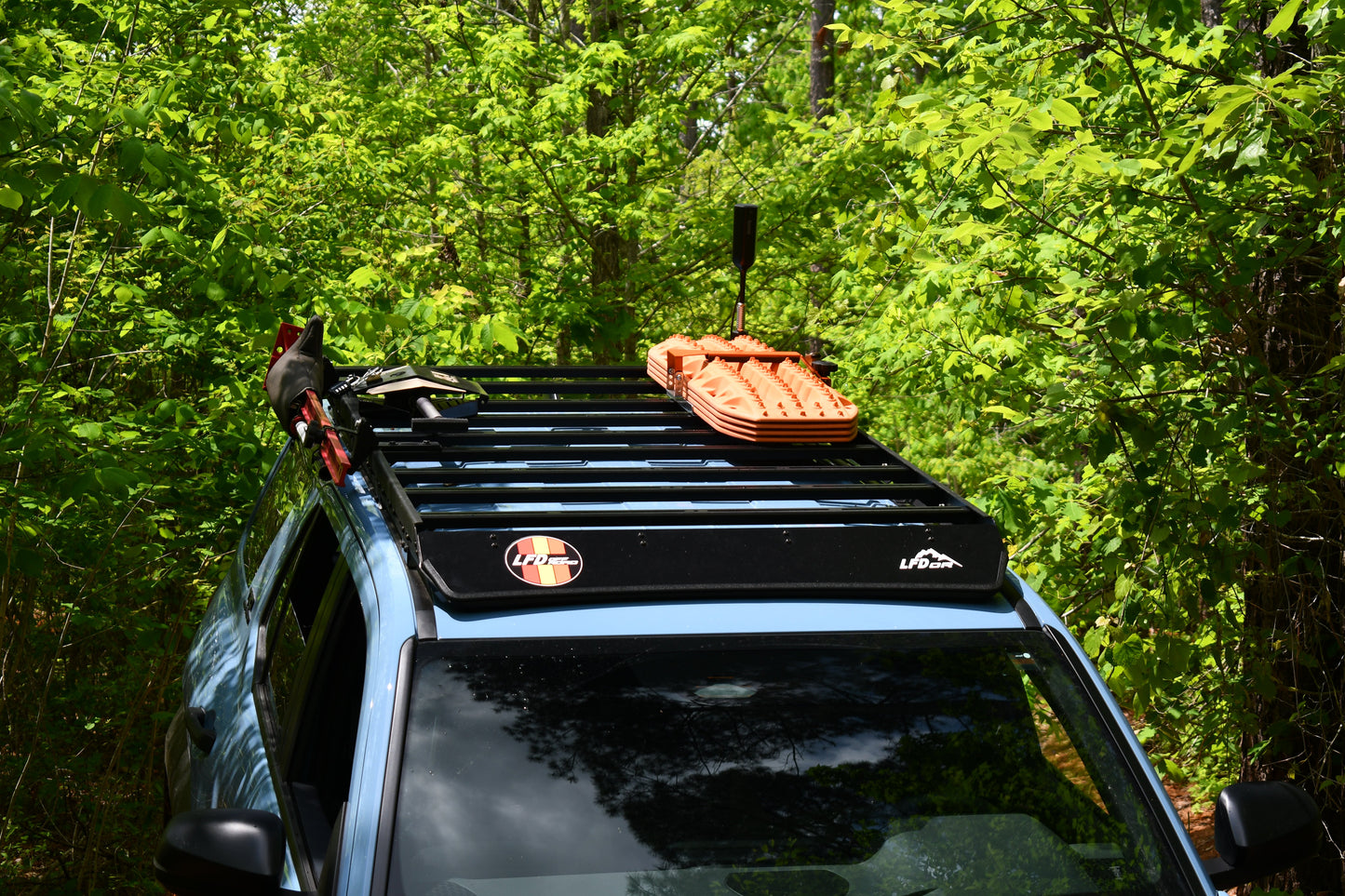 A blue 6th Gen 4Runner with a Ruggedized Low Profile Roof Rack carrying orange traction boards and tools is parked on a narrow, overgrown trail amid dense green foliage.