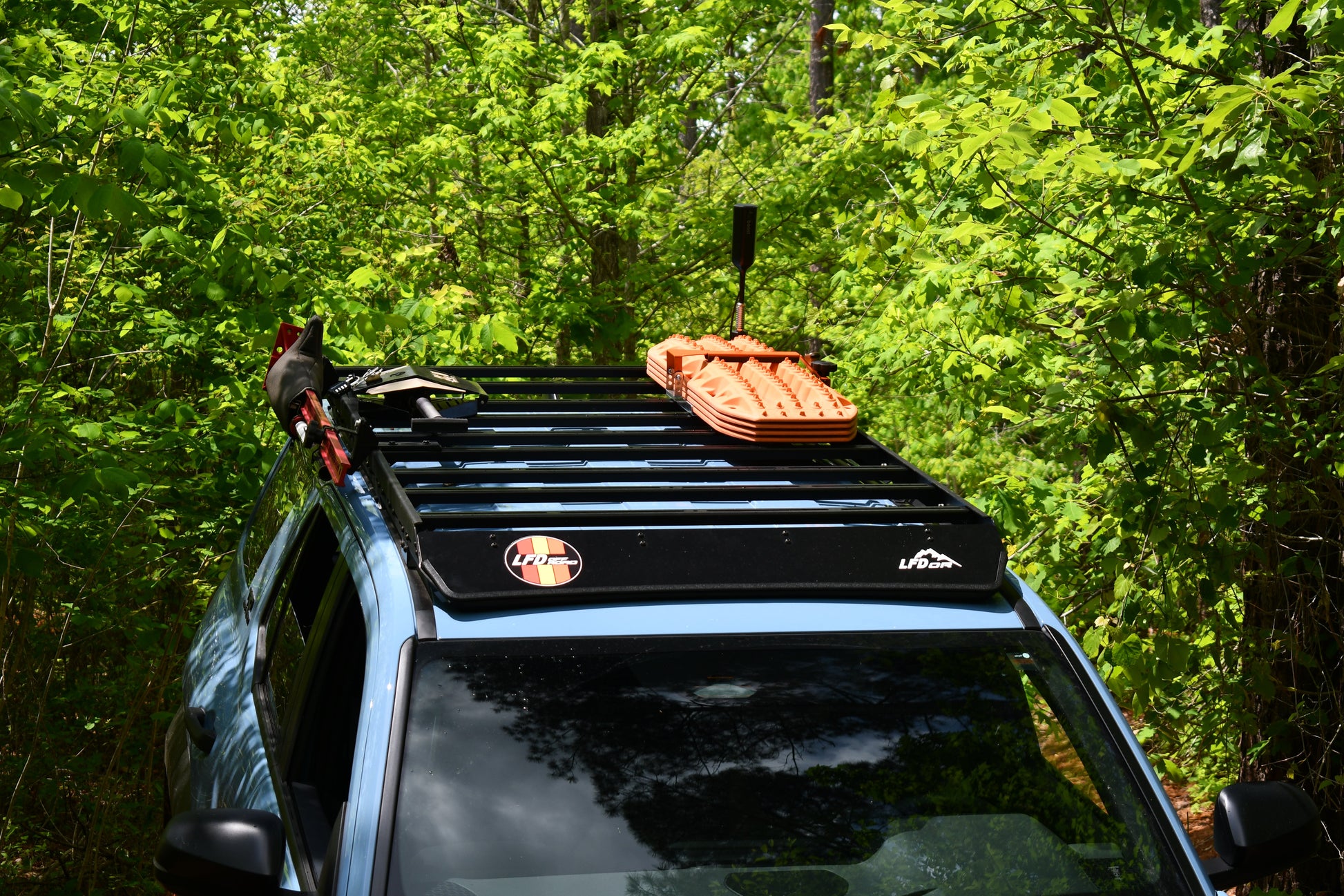 A blue 6th Gen 4Runner with a Ruggedized Low Profile Roof Rack carrying orange traction boards and tools is parked on a narrow, overgrown trail amid dense green foliage.