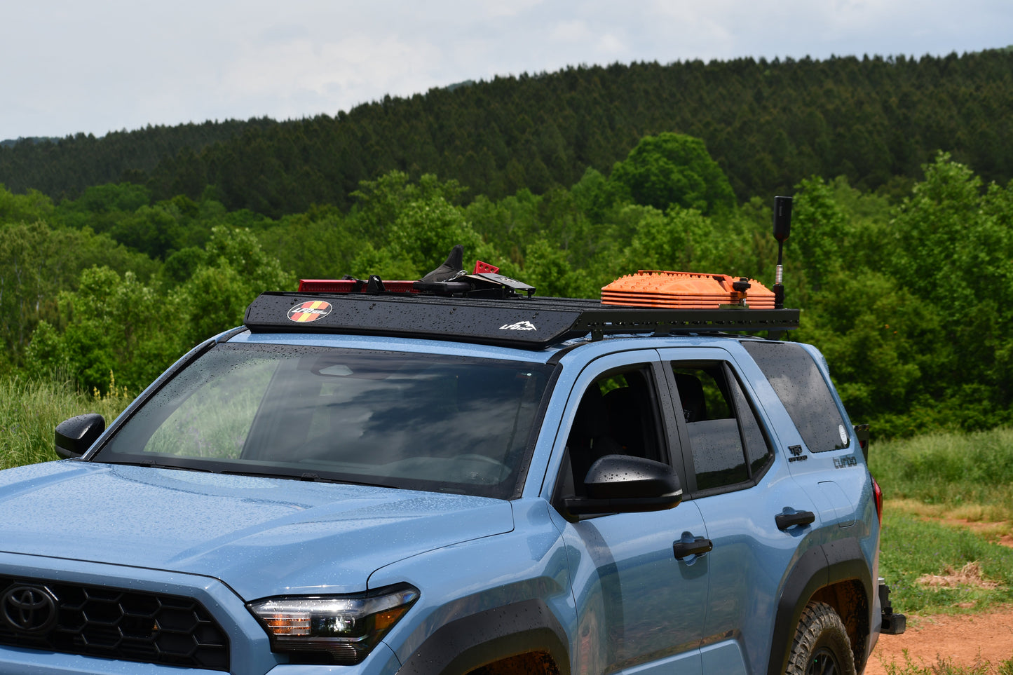 A light blue SUV with a Ruggedized Low Profile Roof Rack - 6th Gen 4Runner, carrying gear like an orange traction board and a shovel, is parked on a dirt road amid green trees and forested hills.