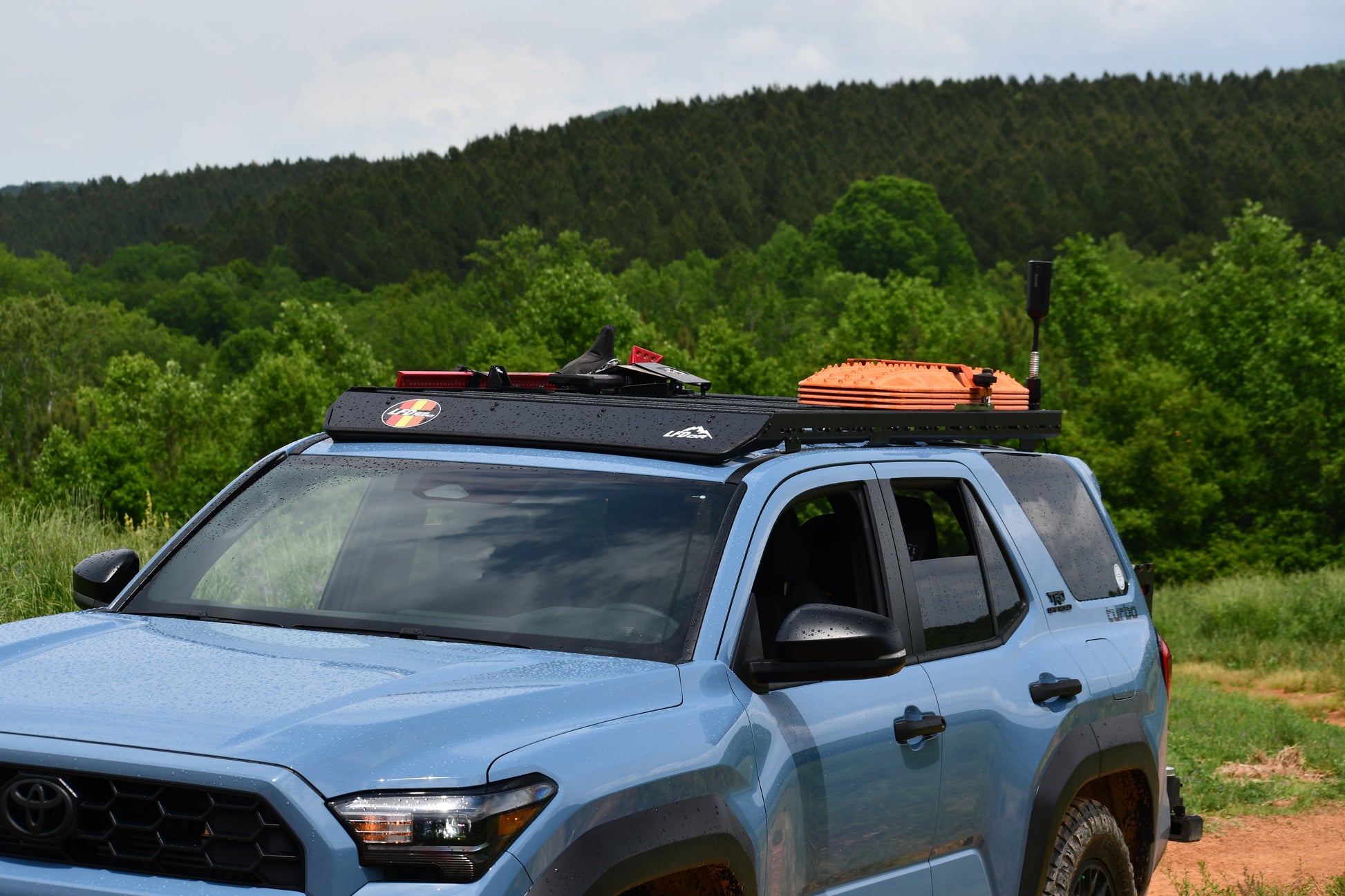 A light blue SUV with a Ruggedized Low Profile Roof Rack - 6th Gen 4Runner, carrying gear like an orange traction board and a shovel, is parked on a dirt road amid green trees and forested hills.