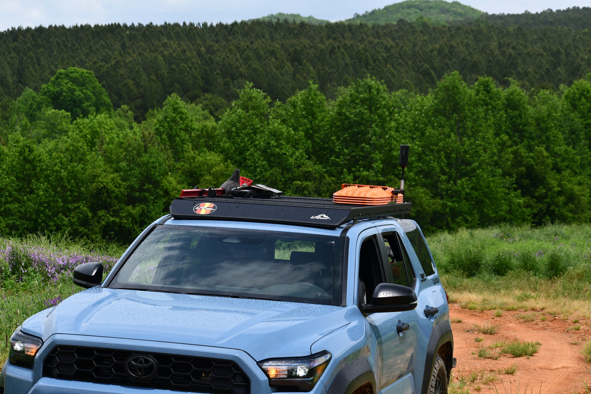 A 6th Gen 4Runner with a Ruggedized Low Profile Roof Rack, loaded with gear and tools, is parked on a dirt road surrounded by green trees and hills beneath a partly cloudy sky.