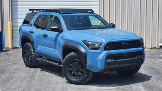 A blue Toyota 4Runner SUV with black wheels is parked in front of a beige industrial building, featuring the Ruggedized Low Profile Roof Rack for the 6th Gen 4Runner.