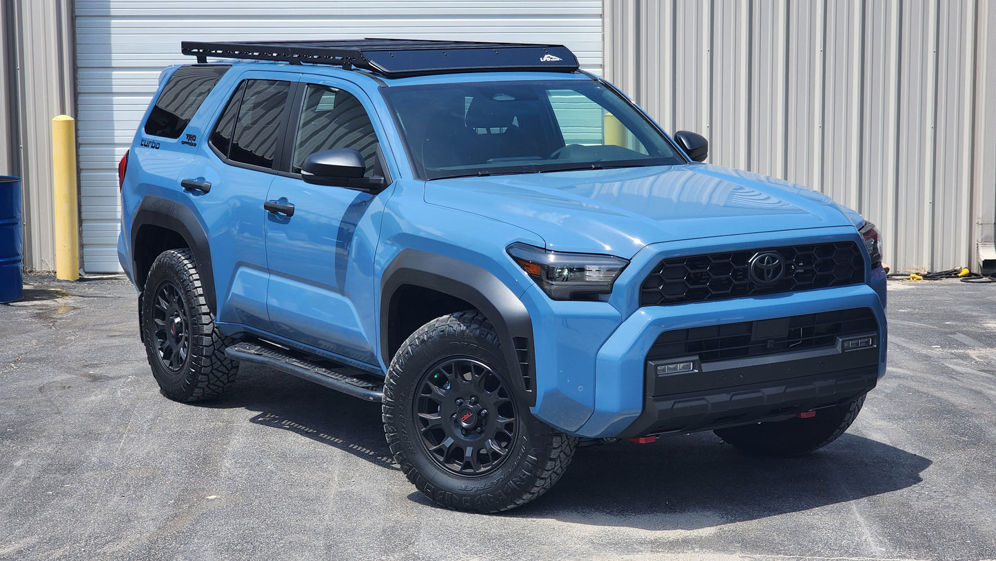 A blue Toyota 4Runner SUV with black wheels and a Ruggedized Low Profile Roof Rack - 6th Gen 4Runner is parked on a paved surface in front of a metal building with a roll-up garage door.