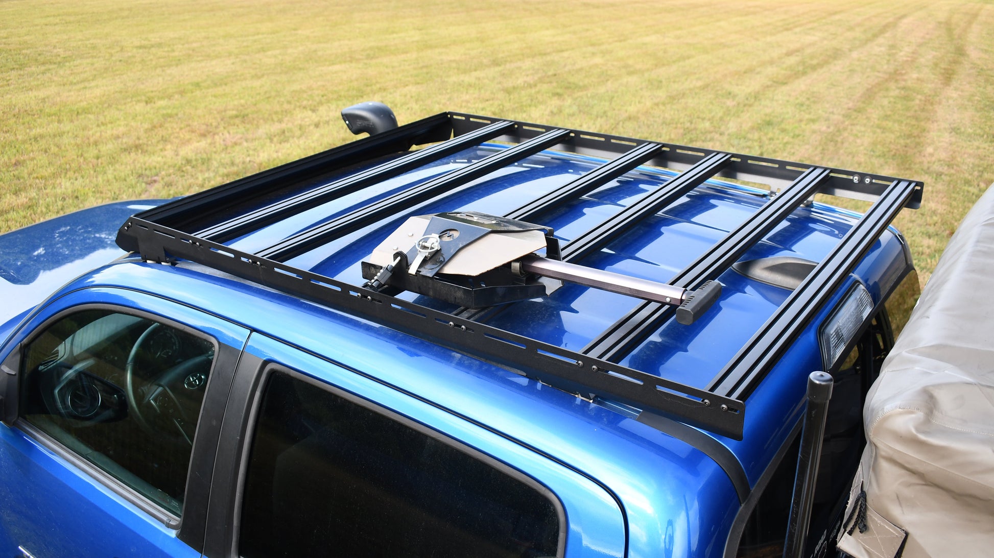 A blue 2nd/3rd Gen Tacoma with a Low Profile Extrusion Rack featuring aluminum crossbars and an awning bracket. The truck, parked on grass, has part of the bed covered by a light-colored tarp.
