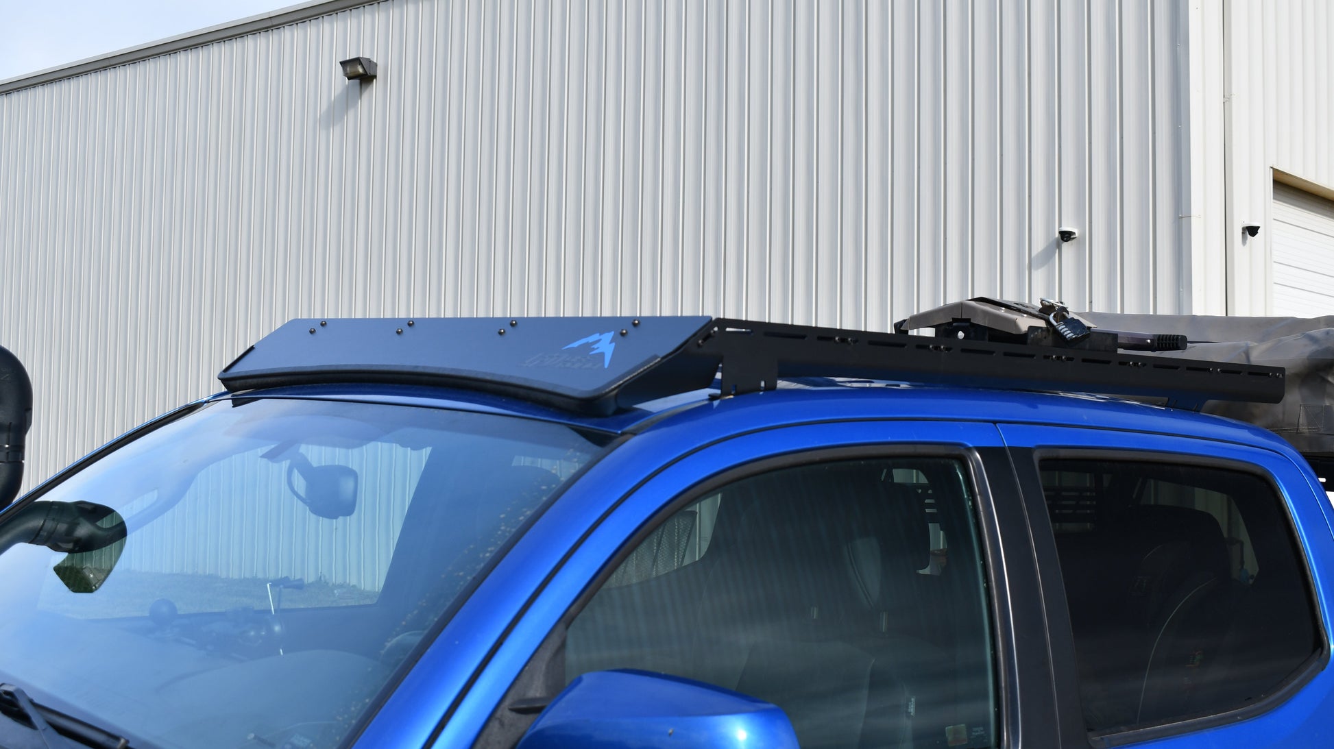 A blue Tacoma with the Low Profile Extrusion Rack (2nd & 3rd Gen) mounted on top, parked beside a corrugated metal building with a white garage door.