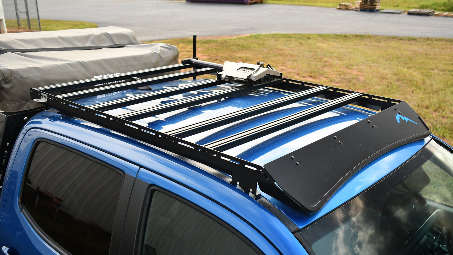 A Low Profile Extrusion Rack - 2nd and 3rd Gen Tacoma is mounted on a blue off-road vehicle with a covered cargo area, parked on grass with industrial pallets visible in the background.