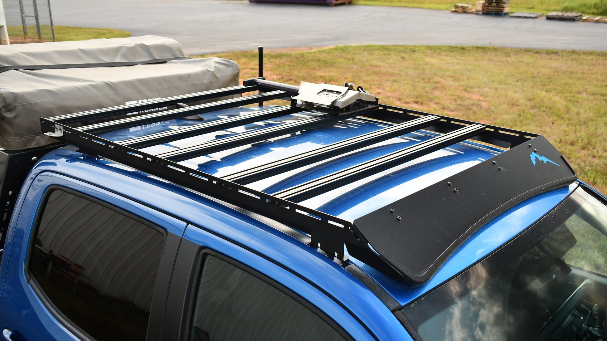 A Low Profile Extrusion Rack - 2nd and 3rd Gen Tacoma is mounted on a blue off-road vehicle with a covered cargo area, parked on grass with industrial pallets visible in the background.