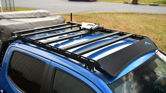 A Low Profile Extrusion Rack - 2nd and 3rd Gen Tacoma is mounted on a blue off-road vehicle with a covered cargo area, parked on grass with industrial pallets visible in the background.