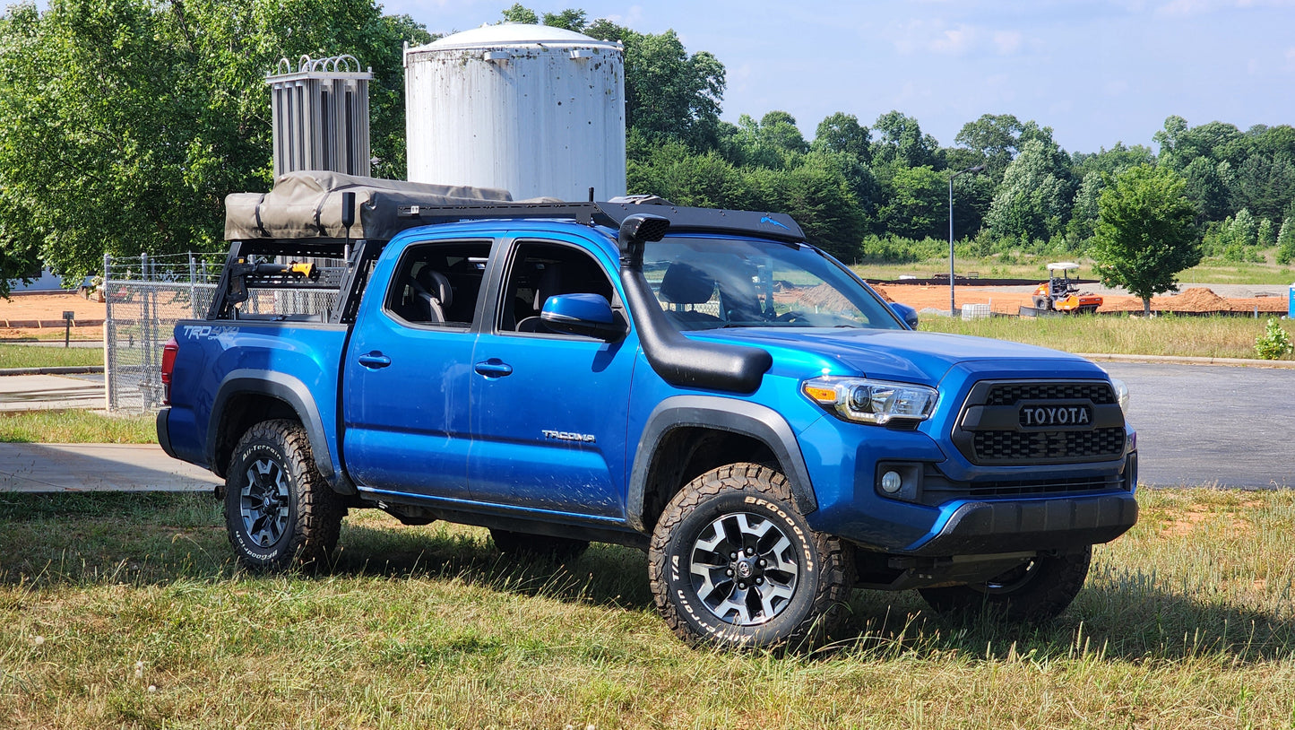A blue Toyota Tacoma TRD Off-Road with off-road tires, a snorkel, and a Low Profile Extrusion Rack - 2nd and 3rd Gen Tacoma carries camping gear, parked on grass near a road with trees in the background.